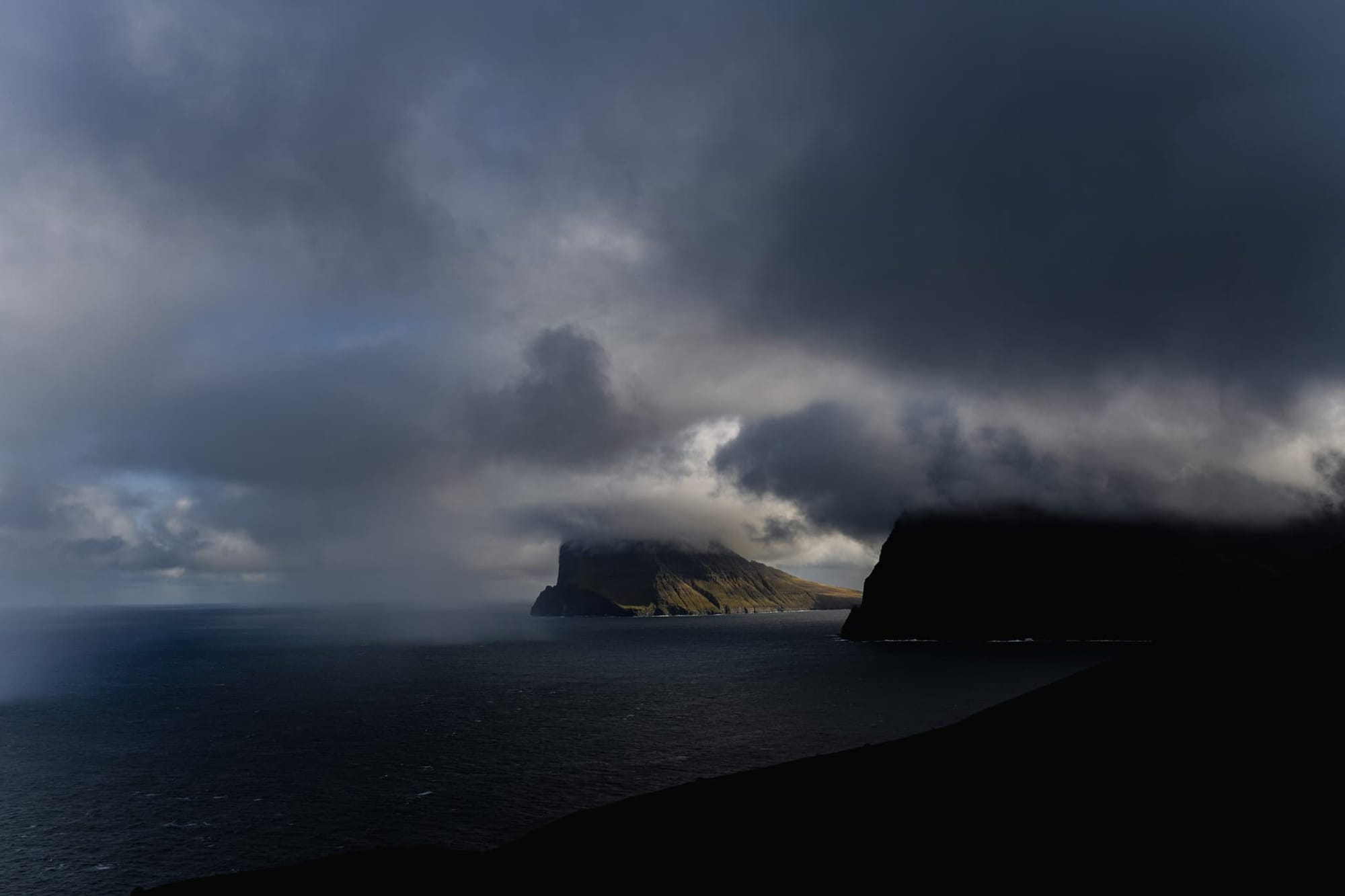 Dramatic storm clouds embrace rugged coastal cliffs in this moody seascape from the Faroe Islands, masterfully captured by elopement photographer Sturmsucht