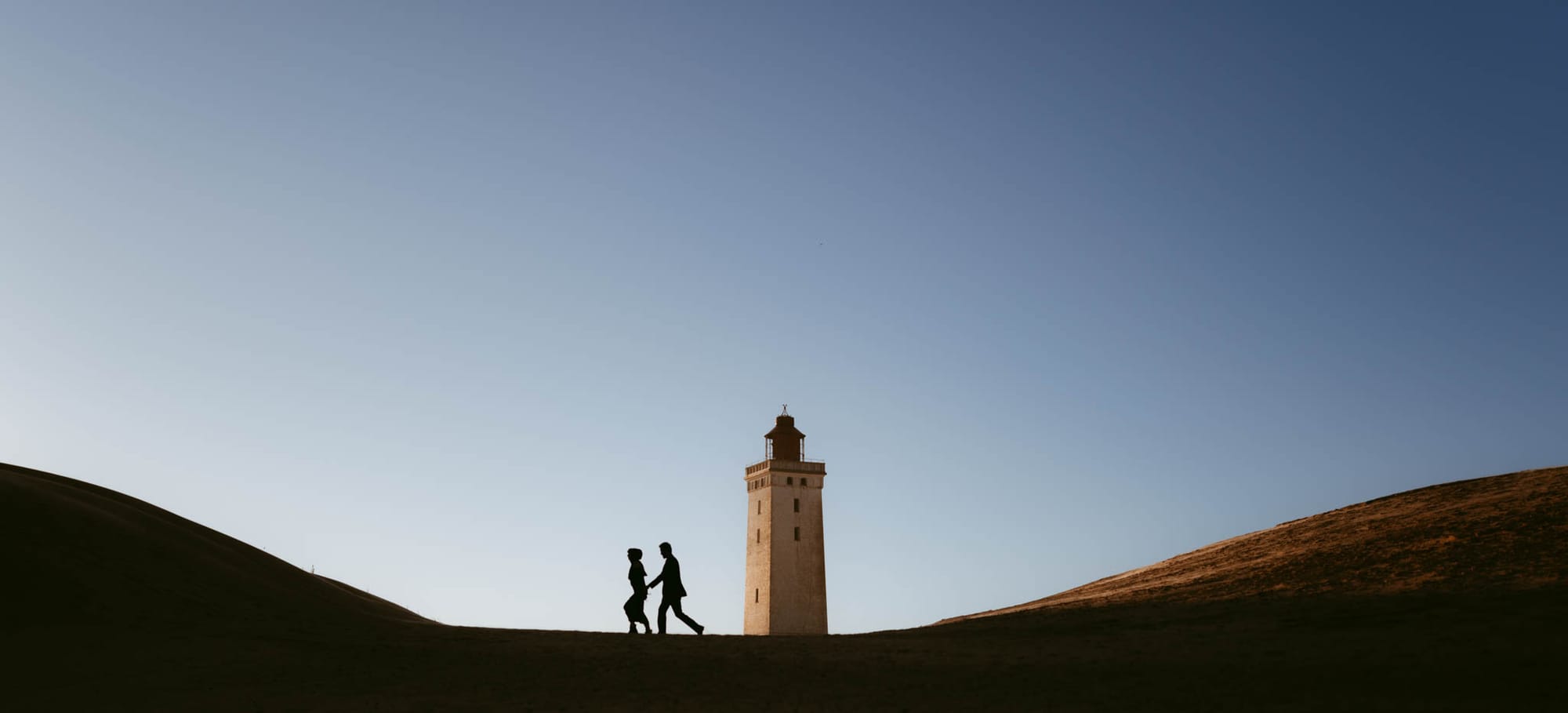 A couple's silhouette against a blue sky as they walk hand in hand towards a lighthouse, symbolizing their elopement in Denmark, photographed by European elopement photographer Sturmsucht