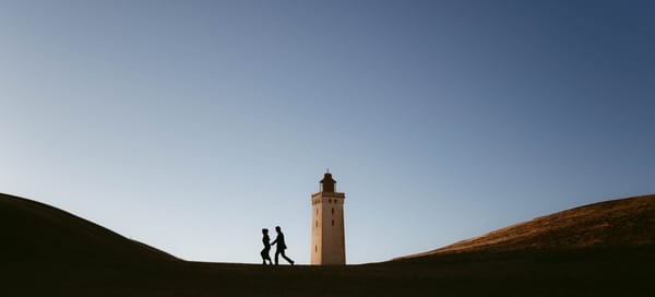 A couple's silhouette against a blue sky as they walk hand in hand towards a lighthouse, symbolizing their elopement in Denmark, photographed by European elopement photographer Sturmsucht